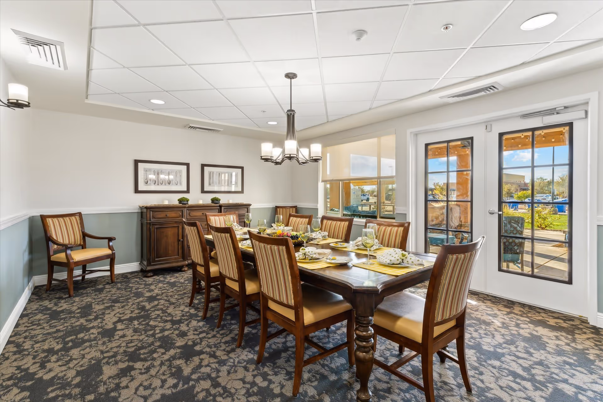 A well-lit dining room with a large wooden dining table set for a meal with plates, glasses, and napkins. The room has eight wooden chairs with striped upholstery around the table, a sideboard against the wall with decorative items, two framed pictures above it, patterned carpet, and double glass doors leading outside to a patio area with greenery visible through the windows.