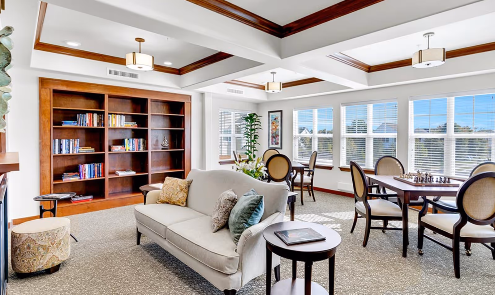 Bright common room with a sofa, side tables, bookshelf, and tables with chairs by large windows under a coffered ceiling.
