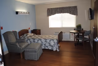 A senior living facility bedroom with a single bed covered in a floral bedspread, a cushioned armchair with an ottoman, a small wooden table with two chairs, a window with blinds and a valance, a wall-mounted TV, and wooden flooring.