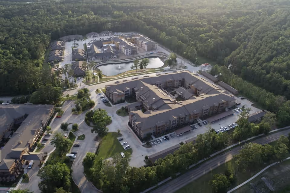 Aerial view of a senior living facility named Watercrest at Kingwood, showing multiple large buildings arranged around a central pond with a fountain, surrounded by dense forest and parking areas.