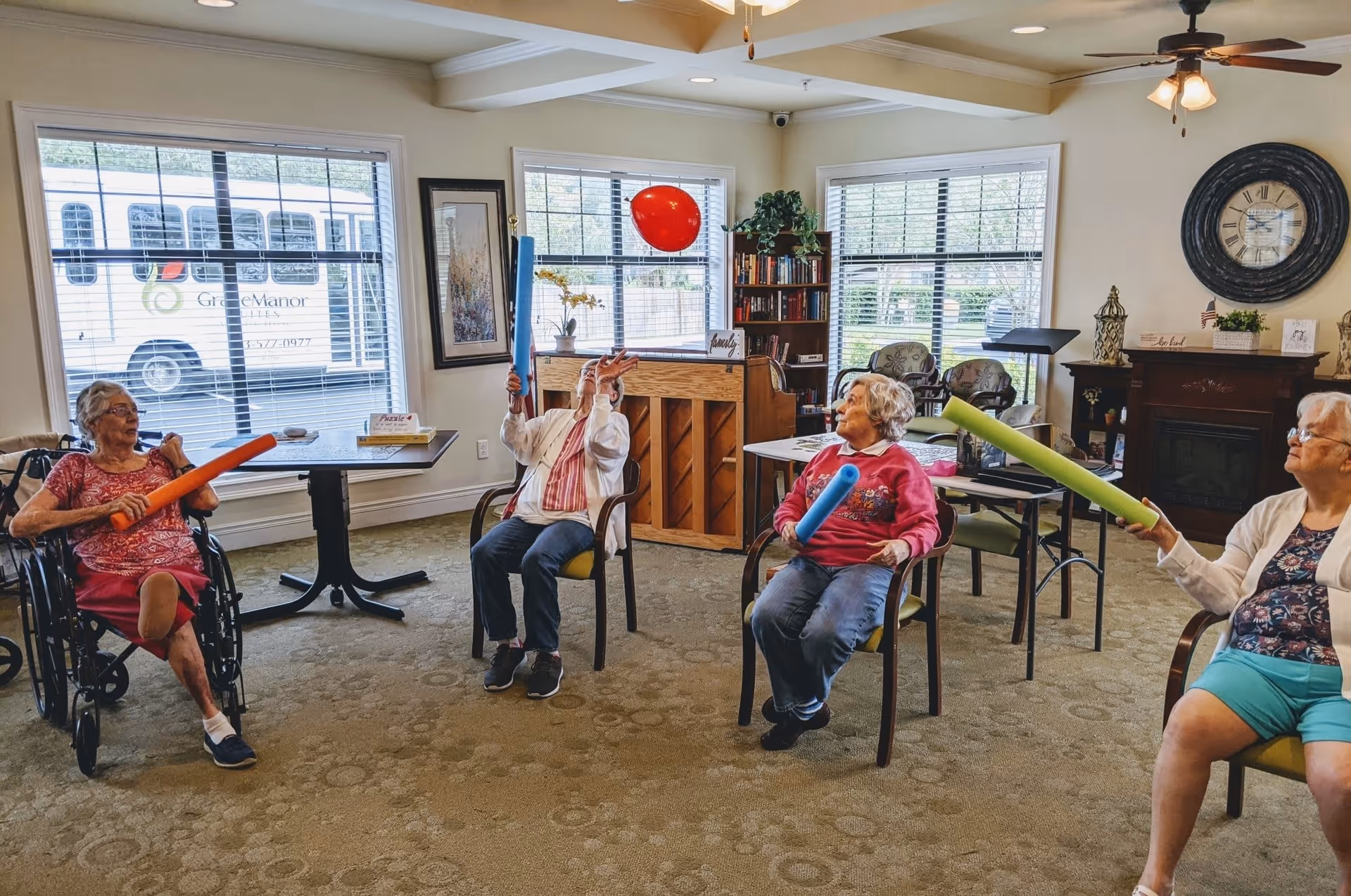 Four elderly women seated in a common room at Grace Manor Suites, each holding colorful foam noodles and playing with a red balloon. The room has large windows with blinds, a piano, bookshelves, a clock on the wall, and comfortable chairs arranged in a circle.