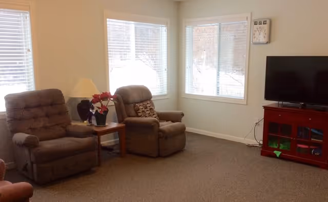 A cozy living room with two brown recliner chairs separated by a wooden side table holding a lamp and a potted plant. There are three large windows with white blinds letting in natural light. A flat-screen TV sits on a red wooden cabinet in the corner, and a wall clock is mounted above the TV.