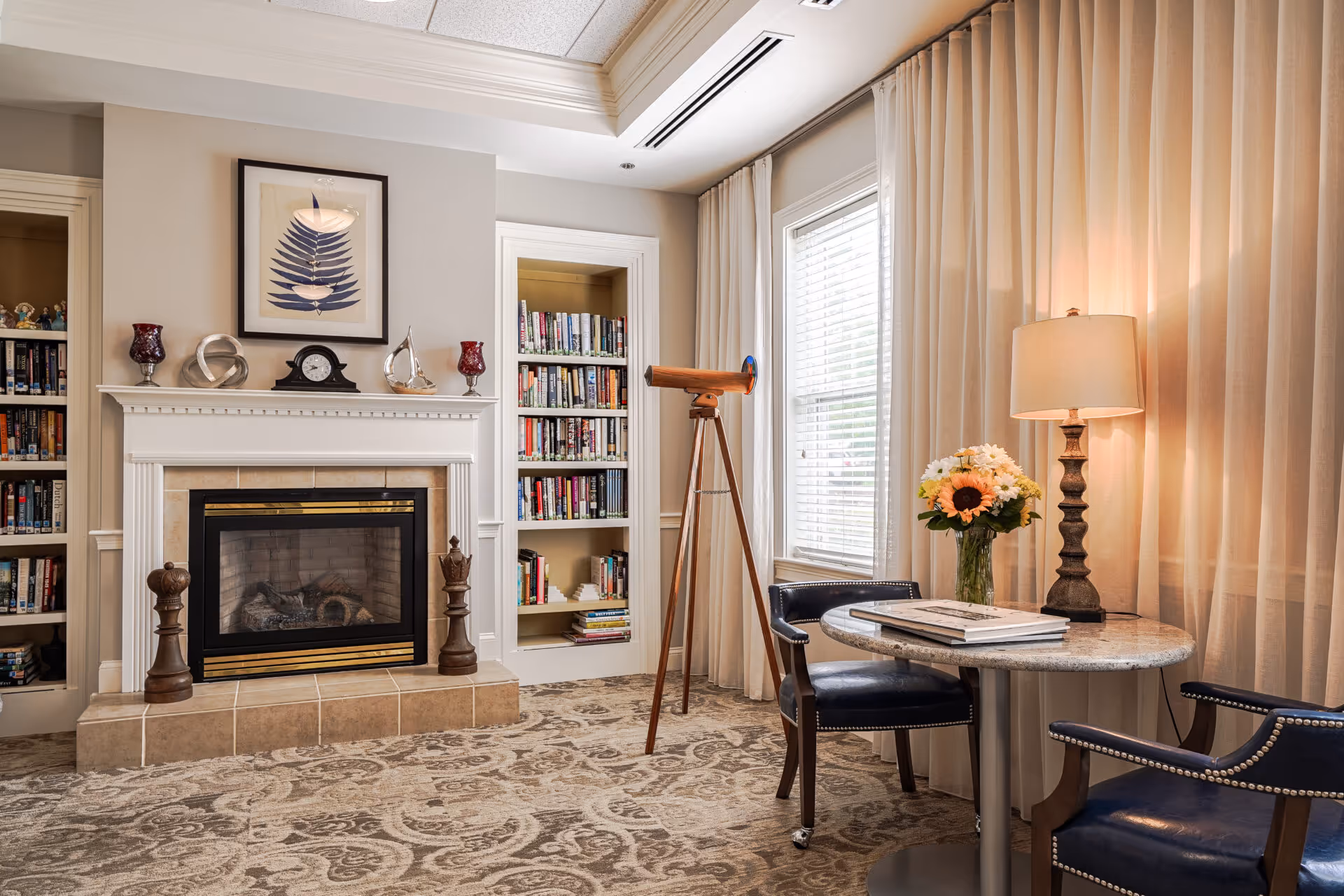 Cozy living room with a fireplace flanked by bookshelves, a telescope by the window, and a small table with chairs and a lamp.
