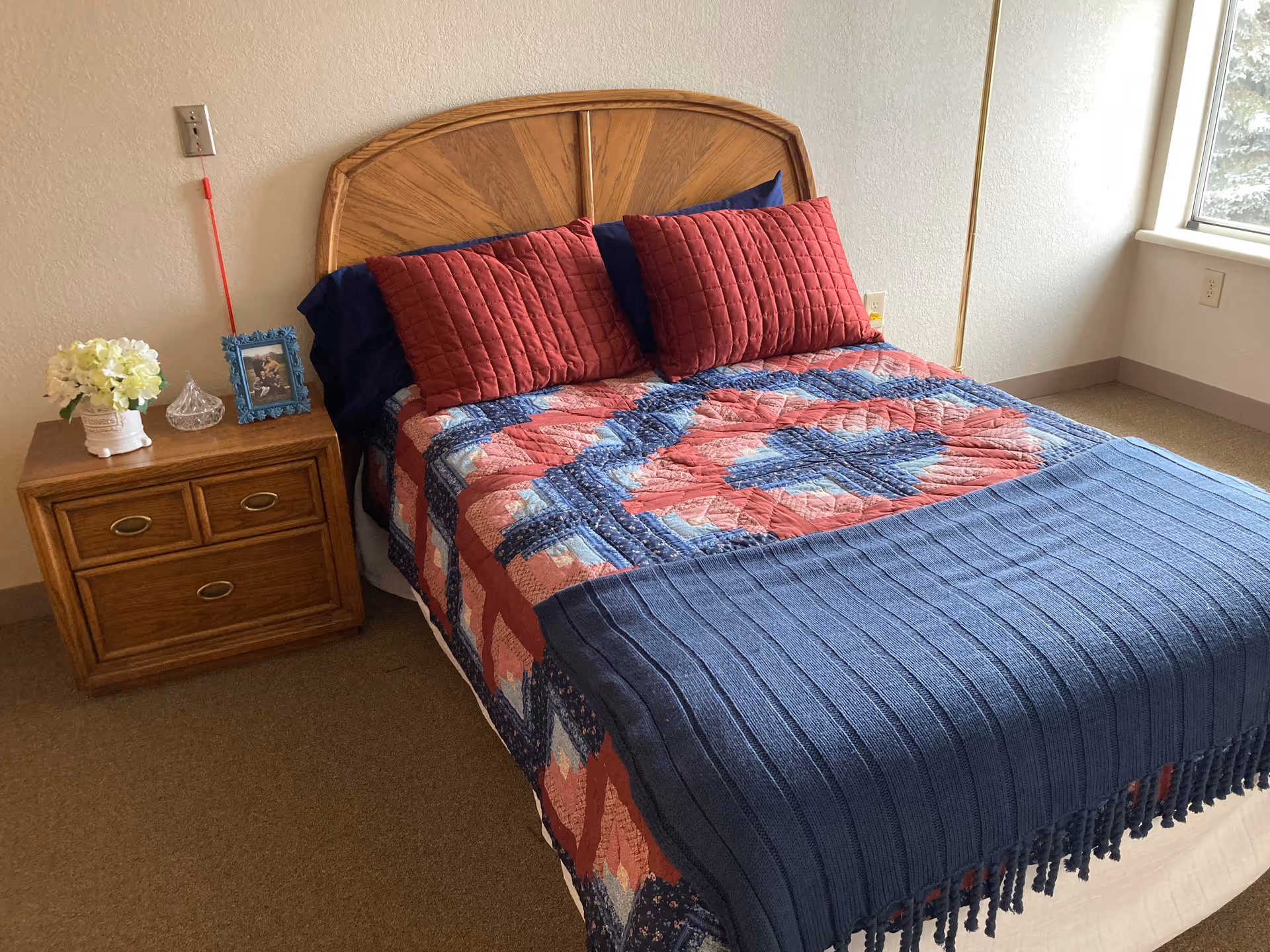 A neatly made bed with a wooden headboard, covered with a colorful quilt featuring red, blue, and white patterns, and two red pillows. Next to the bed is a wooden nightstand with two drawers, holding a small vase of white flowers, a glass container, and a framed photo. A window is visible on the right side, letting in natural light.