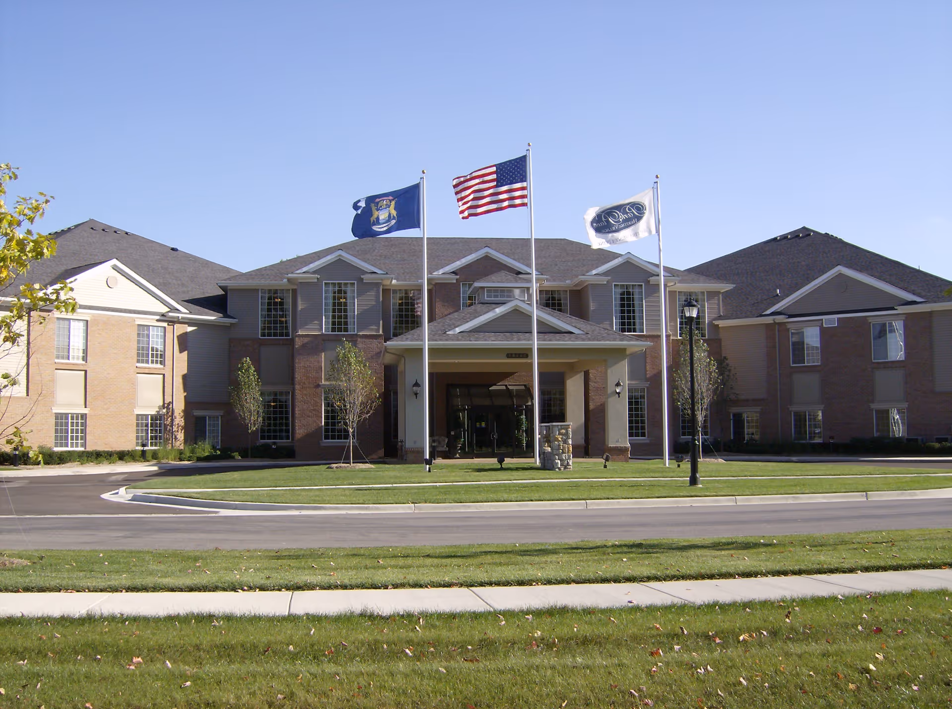 Front exterior view of a two-story senior living facility building with three flagpoles displaying the Michigan state flag, the American flag, and a facility flag. The building has a covered entrance, multiple windows, and a well-maintained lawn with a circular driveway.