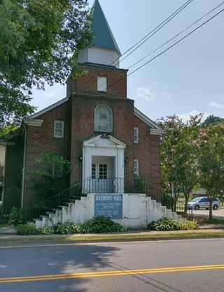 Exterior view of Harmony Hall Assisted Living, a brick building with a tall steeple-like structure, white entrance doors, and a sign in front. Trees and parked cars are visible around the building.