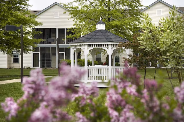 White wooden gazebo in a grassy courtyard with pink flowering shrubs in the foreground and a multi-unit building behind it.