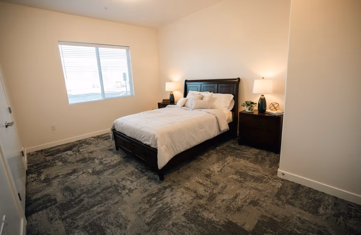 A neatly made bed with white bedding and multiple pillows in a bedroom with beige walls and a large window with blinds. There are two dark wooden nightstands on either side of the bed, each with a lamp and decorative items. The floor is covered with a patterned carpet.