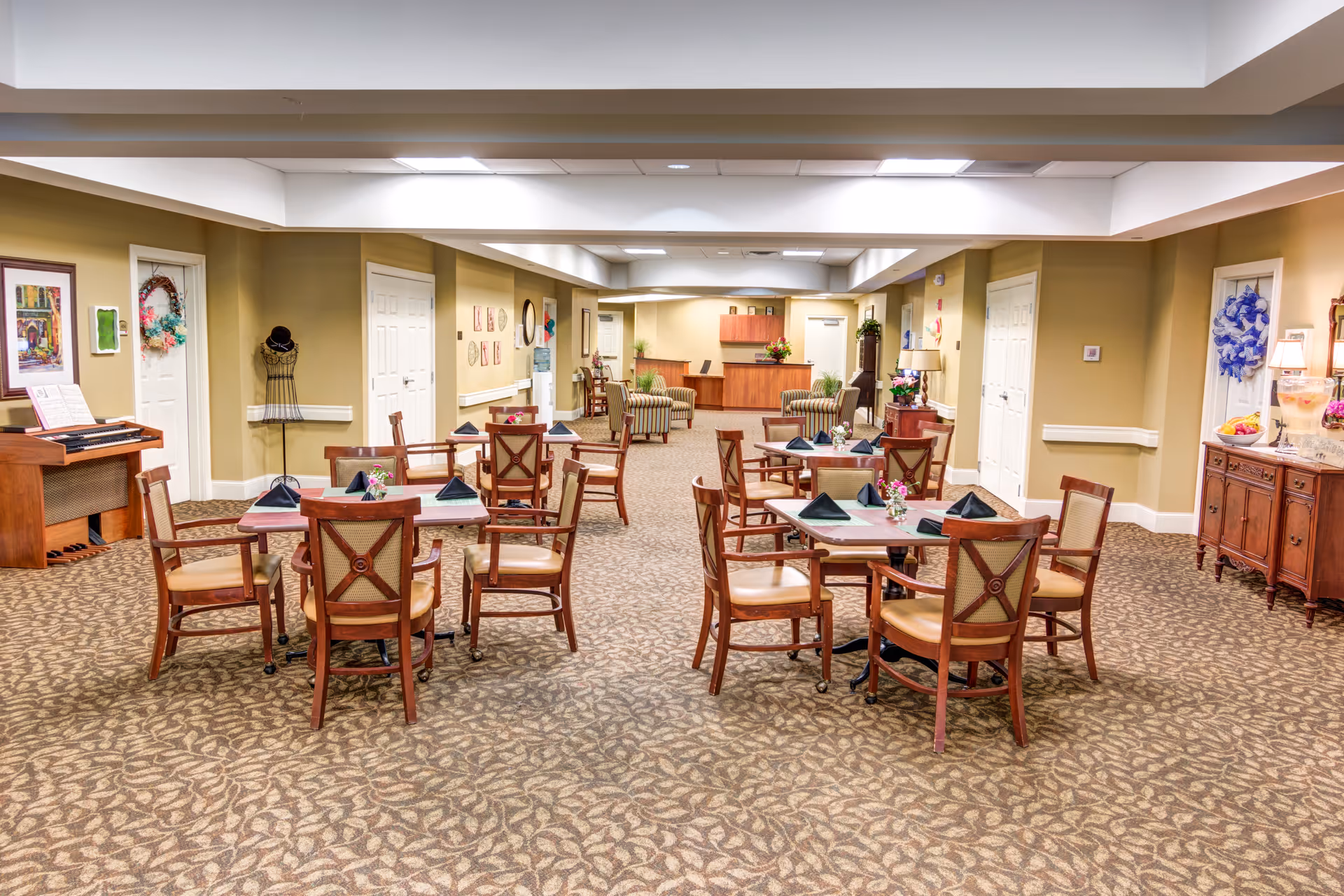 A spacious dining room in a senior living facility with several wooden tables and chairs arranged neatly. Each table has a small floral centerpiece and folded black napkins. The room features beige walls, patterned carpet, and soft lighting. In the background, there is a reception desk with two armchairs and some plants, along with decorative artwork and a piano on the left side.