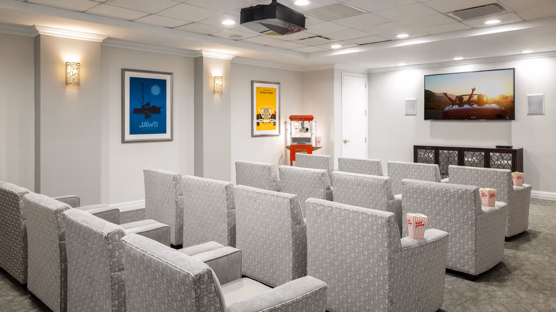A small movie theater room with several rows of cushioned armchairs facing a wall-mounted flat screen TV. The chairs have popcorn containers placed on their armrests. The room has light-colored walls with two framed movie posters and a popcorn machine in the corner.