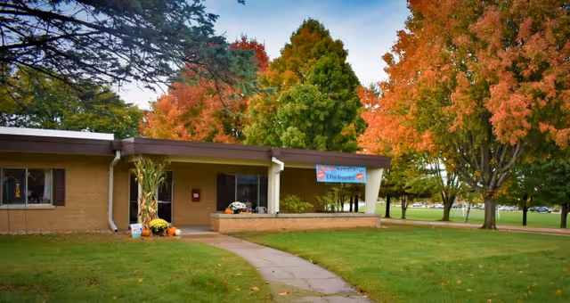Single-story brick building with a covered entrance, surrounded by green grass and trees with autumn foliage. A paved walkway leads to the entrance, and a banner is displayed above the doorway.