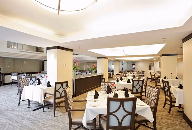 A spacious, well-lit dining room with multiple tables covered in white tablecloths, each set with black napkins, plates, and glassware. The room features patterned chairs, beige walls, carpeted floors, and decorative flower arrangements on a central sideboard.