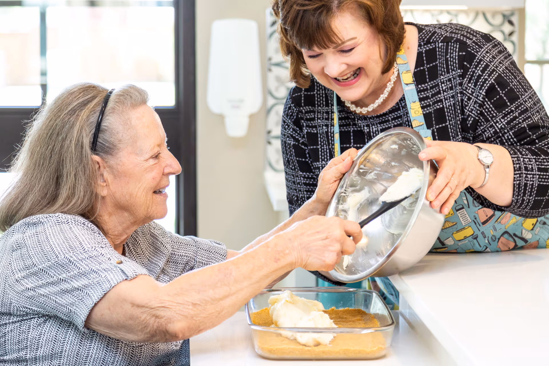 An elderly woman and a caregiver are smiling and preparing a dessert together in a kitchen. The caregiver is holding a mixing bowl and helping the elderly woman spread a creamy mixture onto a crust in a glass baking dish.