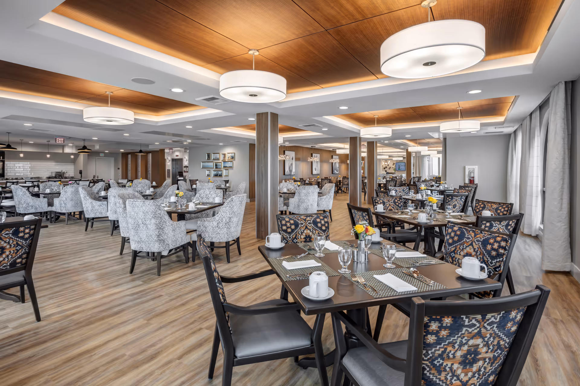 A spacious and well-lit dining room with multiple tables set for meals. The tables have placemats, napkins, cups, glasses, and small flower arrangements. The room features a wood-paneled ceiling with modern circular light fixtures, wood flooring, and large windows with curtains allowing natural light to enter. The chairs have patterned upholstery, and the overall atmosphere is clean and inviting.