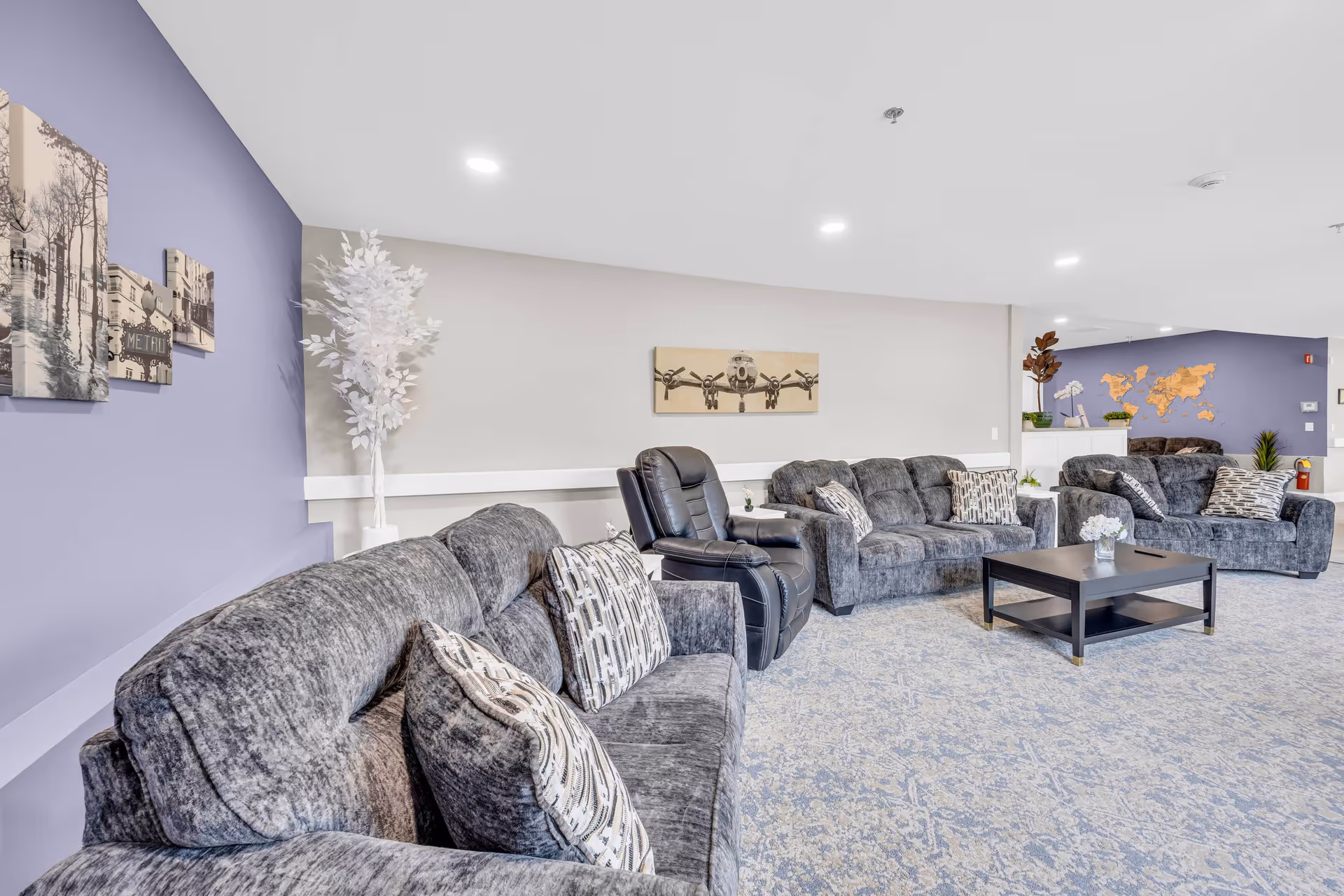 A spacious senior living facility lounge area with multiple gray upholstered sofas and a black recliner chair arranged around a black coffee table. The room features light-colored walls with a purple accent wall adorned with framed artwork and a decorative white plant. The carpet has a subtle blue and gray pattern, and the ceiling has recessed lighting. In the background, there is a wall with a wooden world map and additional seating.