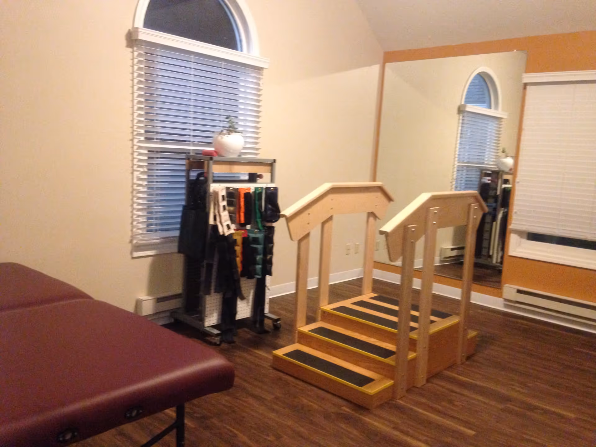 A therapy or rehabilitation room with a wooden exercise staircase with handrails, a padded treatment table, a rack holding various exercise bands, and a large wall mirror reflecting the room. The room has wood flooring, beige walls, and windows with white blinds.
