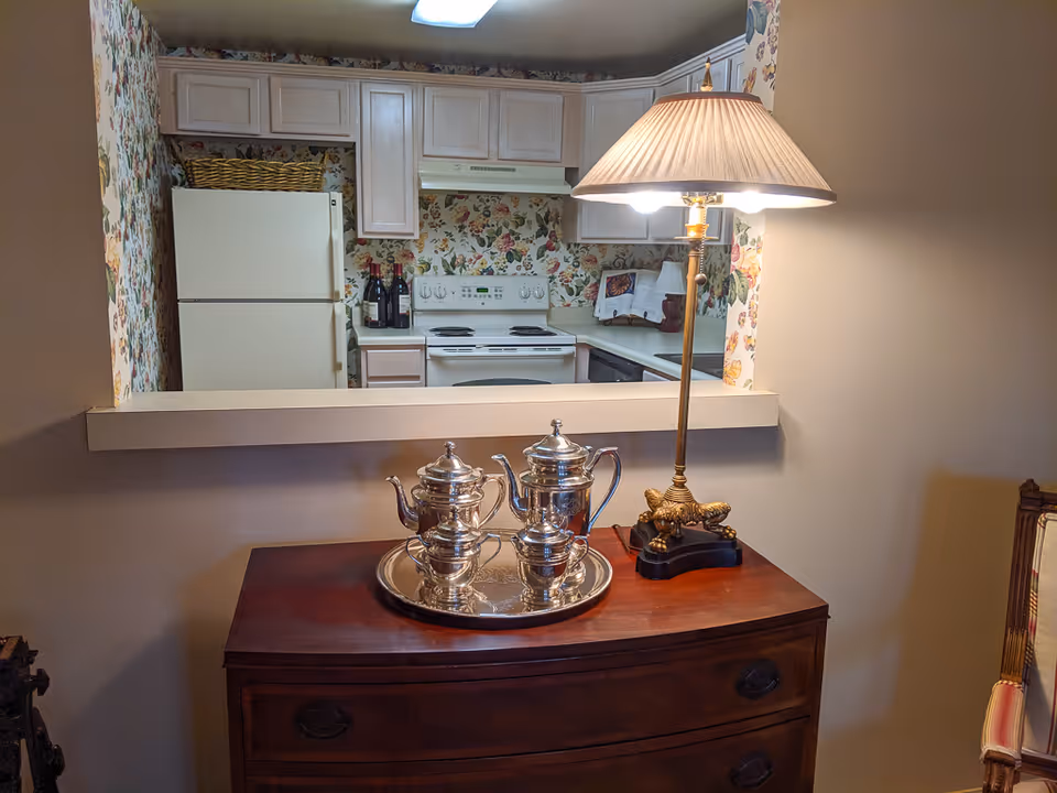 View of a kitchen with floral wallpaper, white cabinets, a white refrigerator, and a white stove. In the foreground, there is a wooden dresser with a silver tea set on a tray and a decorative table lamp with a pleated lampshade. A chair with striped upholstery is partially visible on the right side.