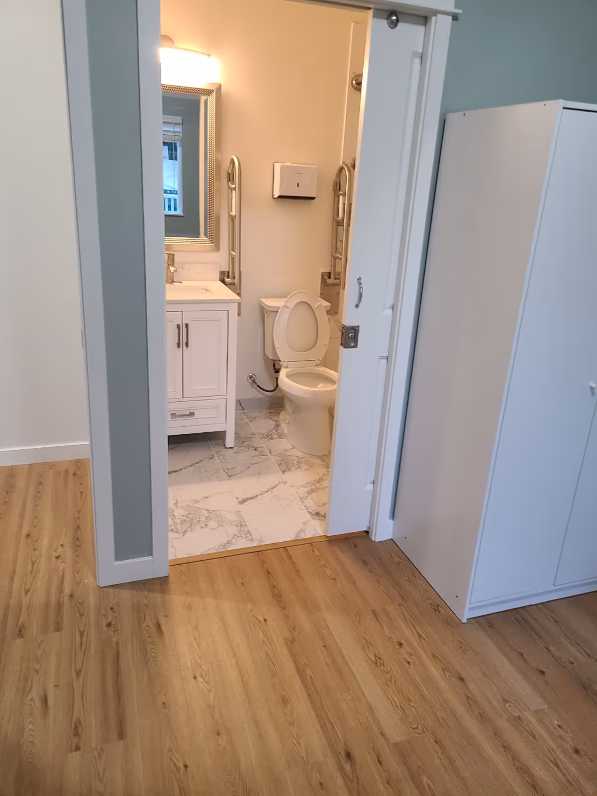Open doorway revealing a bathroom with a toilet, sink vanity, mirror, and grab bars adjacent to a room with wood flooring.