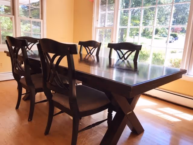 A dining area with a dark wooden rectangular table and six matching chairs with cushioned seats. The room has large windows letting in natural light and a view of greenery outside. The walls are painted a warm yellow color and the floor is wooden.