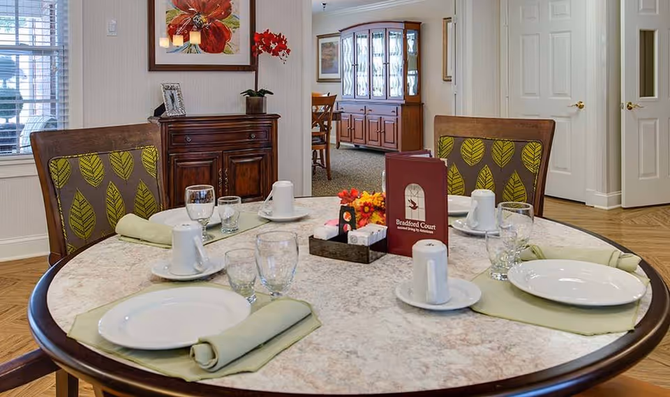 A round dining table set for four with white plates, cups, glasses, and light green napkins. The chairs have a wooden frame with green leaf-patterned upholstery. In the center of the table is a small flower arrangement and a red menu labeled 'Bradford Court'. The room has light-colored walls, wooden flooring, and a wooden cabinet with a framed picture and a red flower pot on top. A dining room with a china cabinet is visible in the background.