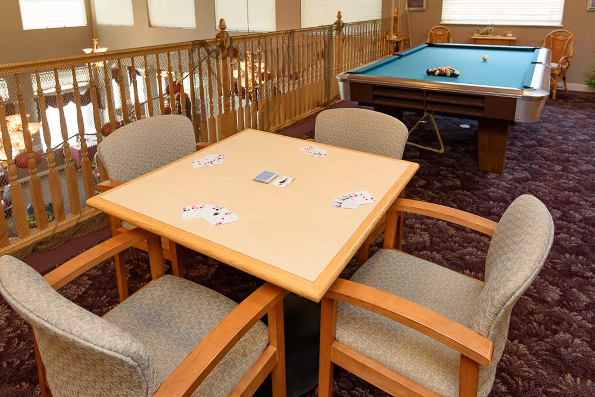 Game room with a card table surrounded by four chairs and a pool table in the background.