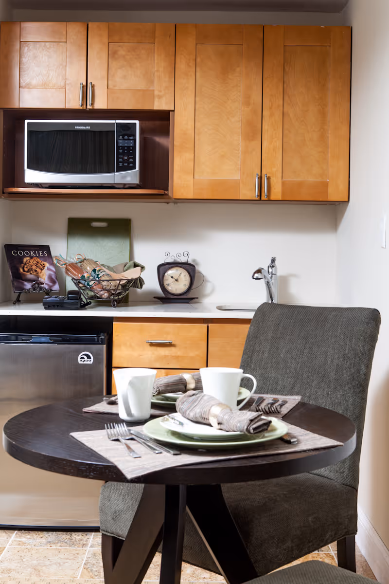 A small kitchen area with wooden cabinets, a microwave, a mini refrigerator, and a countertop with a decorative clock and a basket. In the foreground, there is a round dark wooden table set for two with plates, cups, utensils, and cloth napkins, accompanied by two upholstered chairs.