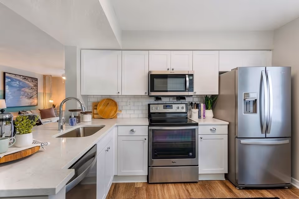 Modern kitchen with white cabinets, stainless steel refrigerator, oven, and microwave. The kitchen has a white countertop with a sink and a few decorative items including a plant and cutting boards. In the background, part of a living room with a large wall art and sofa is visible.