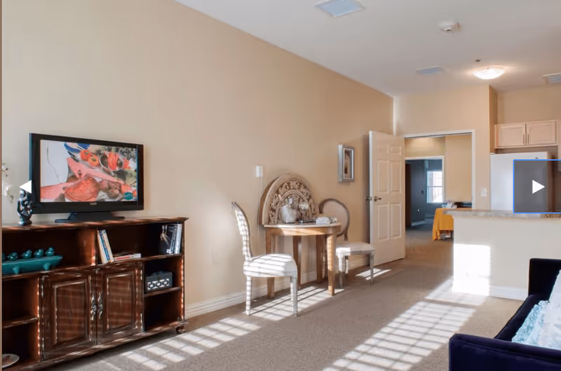 A bright living area with beige walls and carpeted floor. There is a wooden TV stand with a flat-screen TV on top displaying a colorful image. Next to it is a small round wooden table with two striped chairs. An open doorway leads to another room with a bed visible. The kitchen area with light wood cabinets and a refrigerator is partially visible on the right. Sunlight streams through a window, casting shadows on the floor.