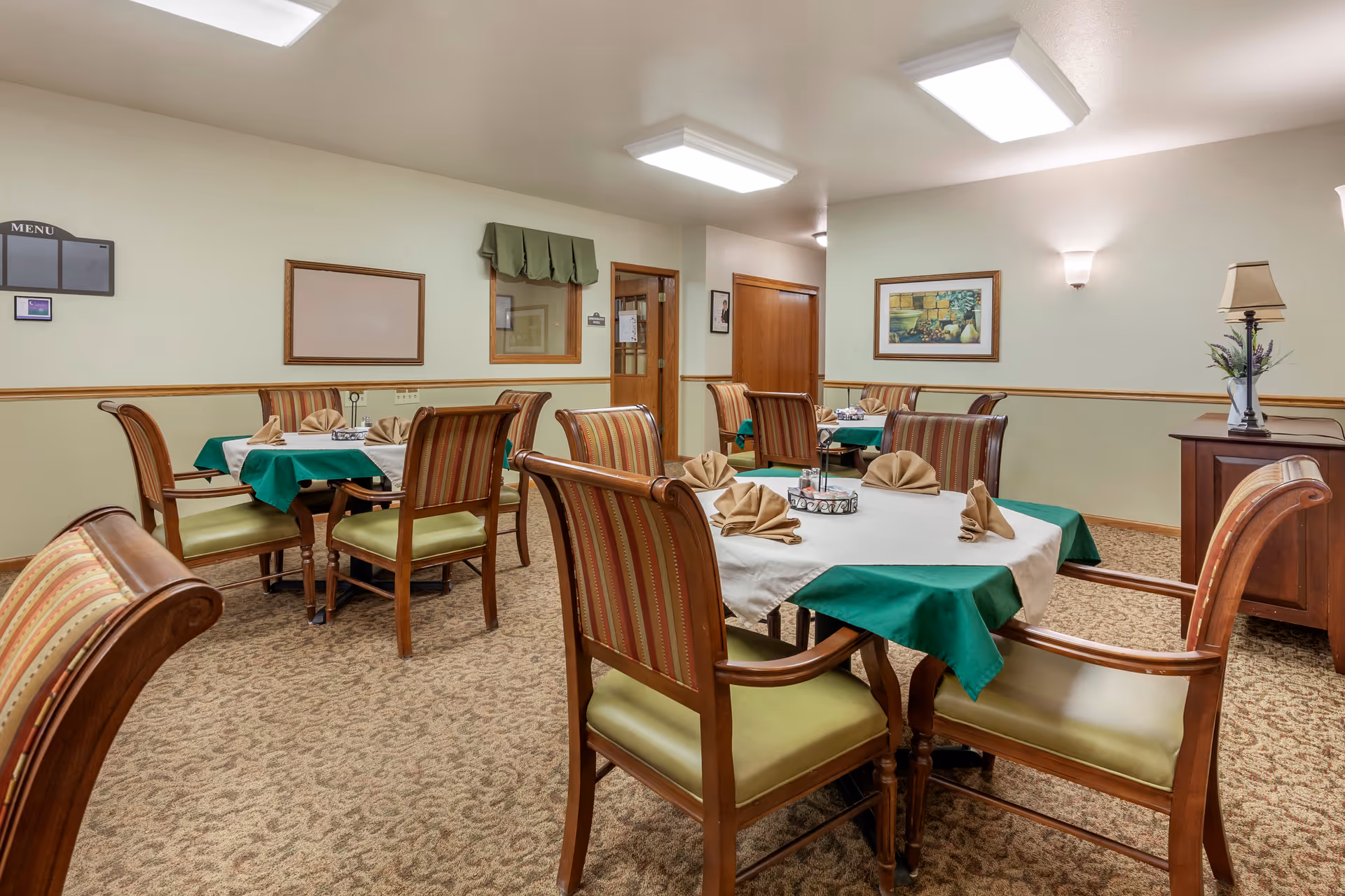 Dining room with several set tables, wooden chairs, green tablecloths and folded napkins in a senior living facility.