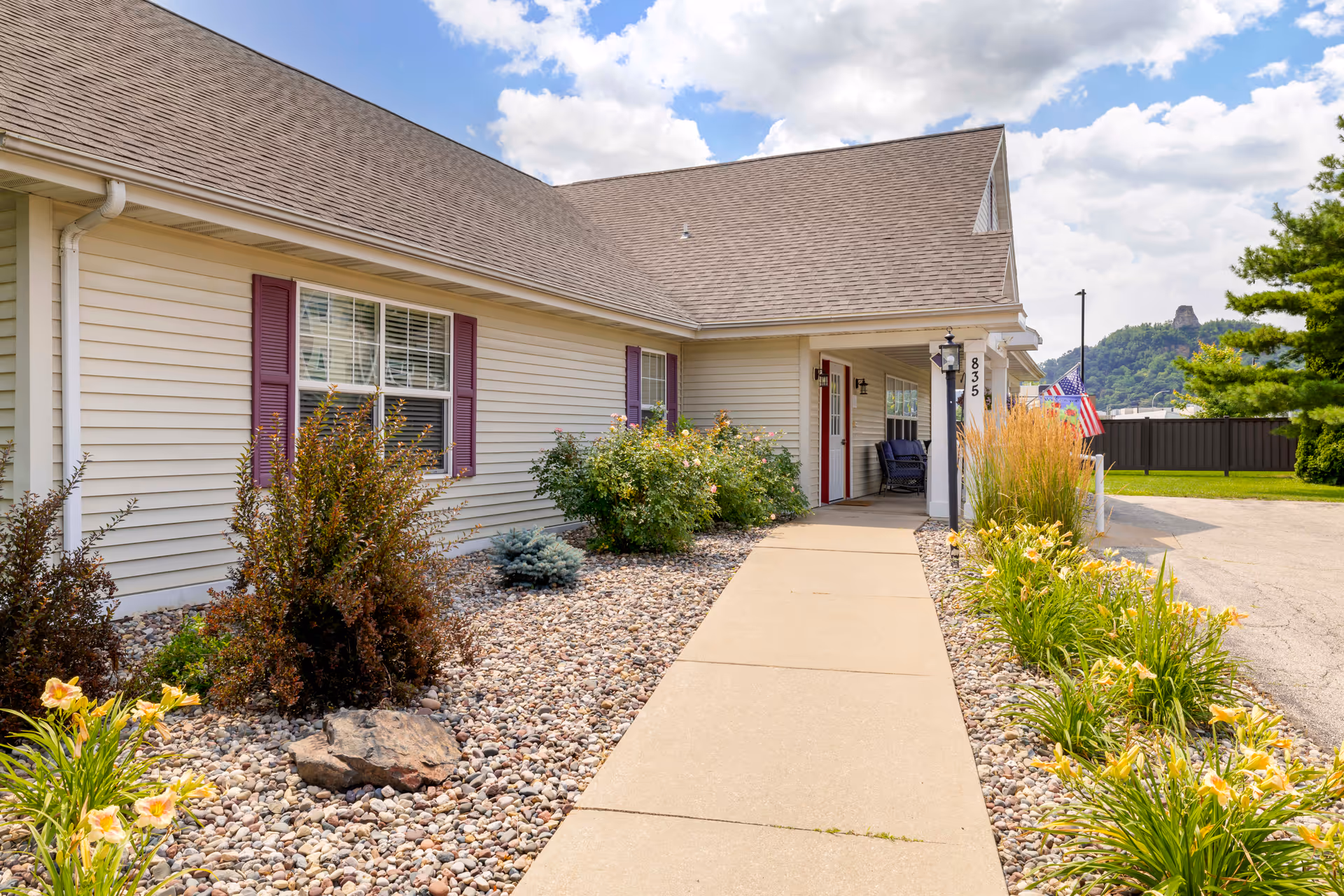 Exterior view of a single-story building with beige siding and purple shutters. A concrete walkway leads to a covered entrance with a red door and two chairs on the porch. The landscaping includes rocks, bushes, and yellow flowers. An American flag is visible near the entrance, and a green hill is in the background under a partly cloudy sky.