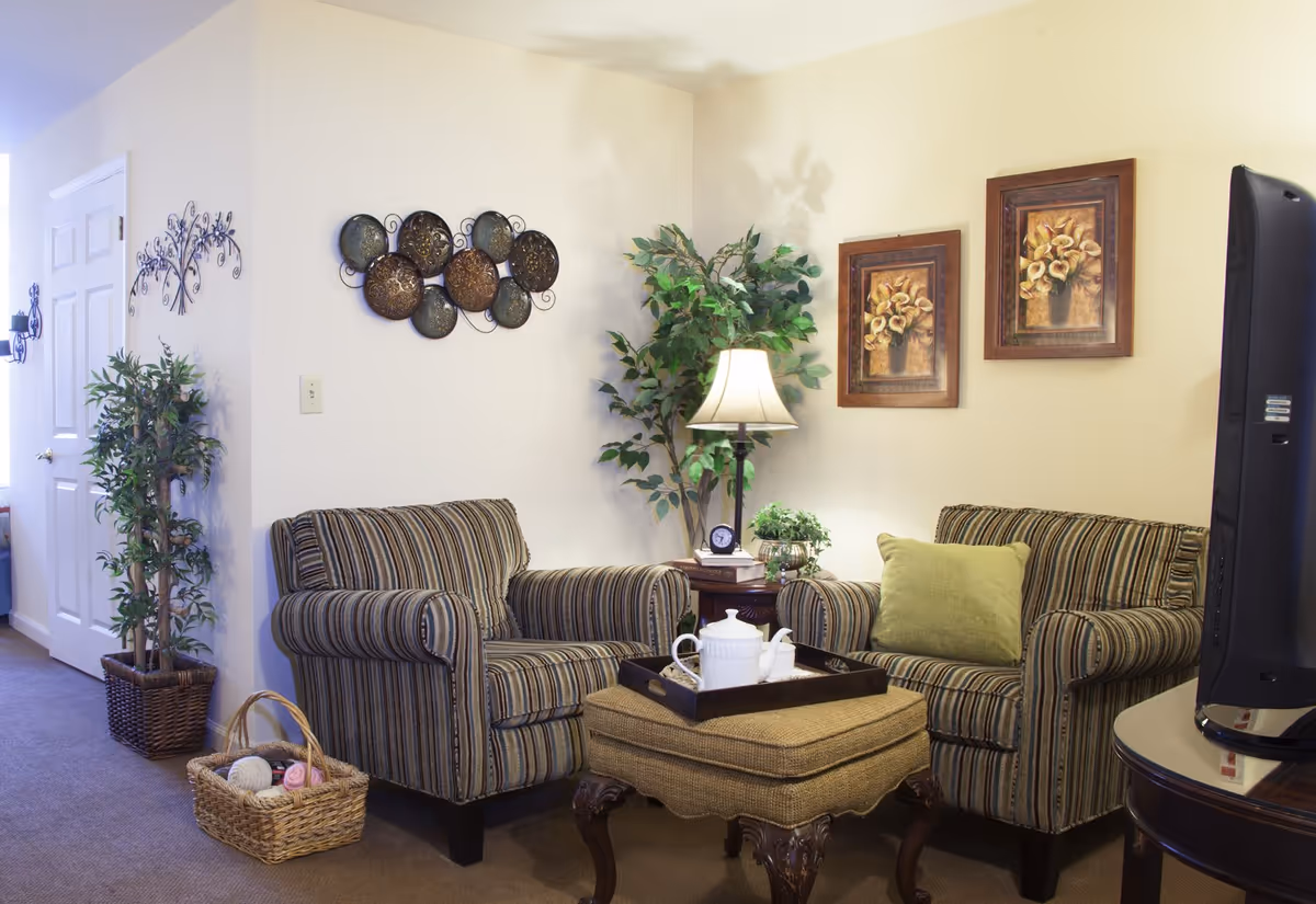 A cozy living room area with two striped armchairs, a cushioned ottoman with a tray holding a teapot and cups, a side table with a lamp, clock, and plant, wall decorations including metal art and framed floral paintings, a potted plant in the corner, and a TV on a round table.