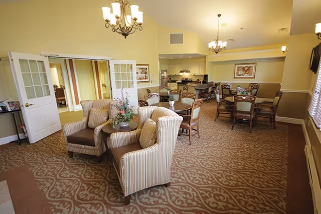 A spacious assisted-living common room with upholstered armchairs, small round tables, a dining area and a kitchenette under chandeliers.