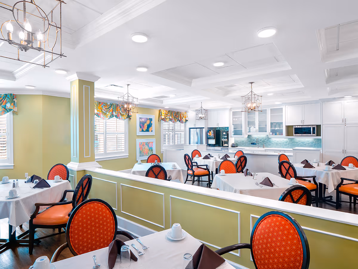Bright and elegant dining room with multiple tables covered in white tablecloths, each set with brown napkins, cups, and silverware. The chairs have orange patterned cushions and dark wooden frames. The room features light green walls, large windows with colorful valances, and a white coffered ceiling with hanging lantern-style light fixtures. In the background, there is a modern kitchen area with white cabinetry, a refrigerator, microwave, and a coffee machine.