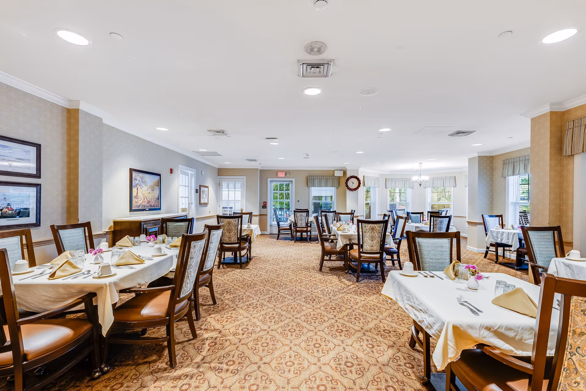 A spacious dining room in a senior living facility with multiple tables covered in white tablecloths, each set with beige napkins, cups, and silverware. The room has patterned carpet, light-colored walls with framed artwork, large windows with valances, and wooden chairs with cushioned seats. A clock is visible on the far wall near the exit door.