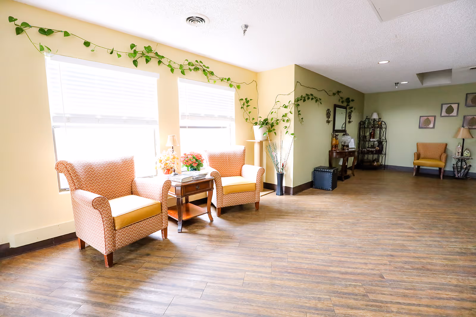 Bright communal seating area with upholstered armchairs, a side table with flowers, potted plants, and decorative shelving along wood flooring.