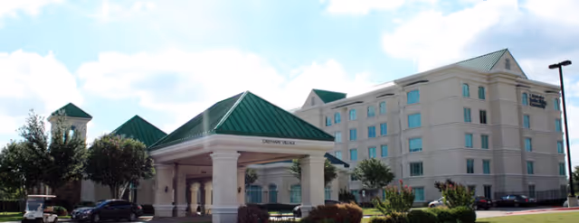 Exterior view of a multi-story senior living facility with a green roof and a covered entrance labeled 'Greenway Village'. Several trees and parked cars are visible around the building under a partly cloudy sky.