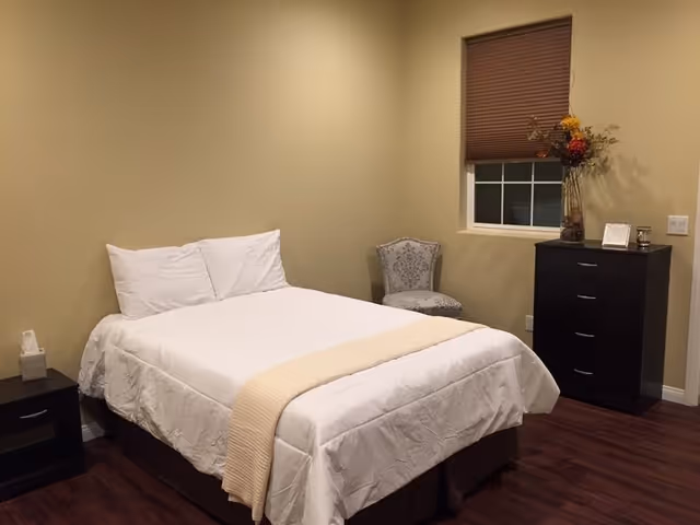 A simple bedroom with a neatly made bed featuring white bedding and a beige throw. There is a small black nightstand with a tissue box on the left side of the bed, a decorative chair with a patterned fabric in the corner, and a black chest of drawers with a vase of flowers and a small framed picture on top. The room has beige walls, a window with a closed brown blind, and dark wood flooring.
