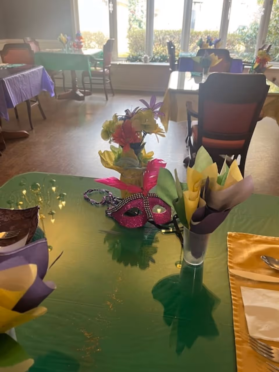 A dining room with tables covered in colorful tablecloths and decorated with artificial flowers and a pink masquerade mask. Chairs are arranged around the tables, and large windows in the background let in natural light with a view of greenery outside.