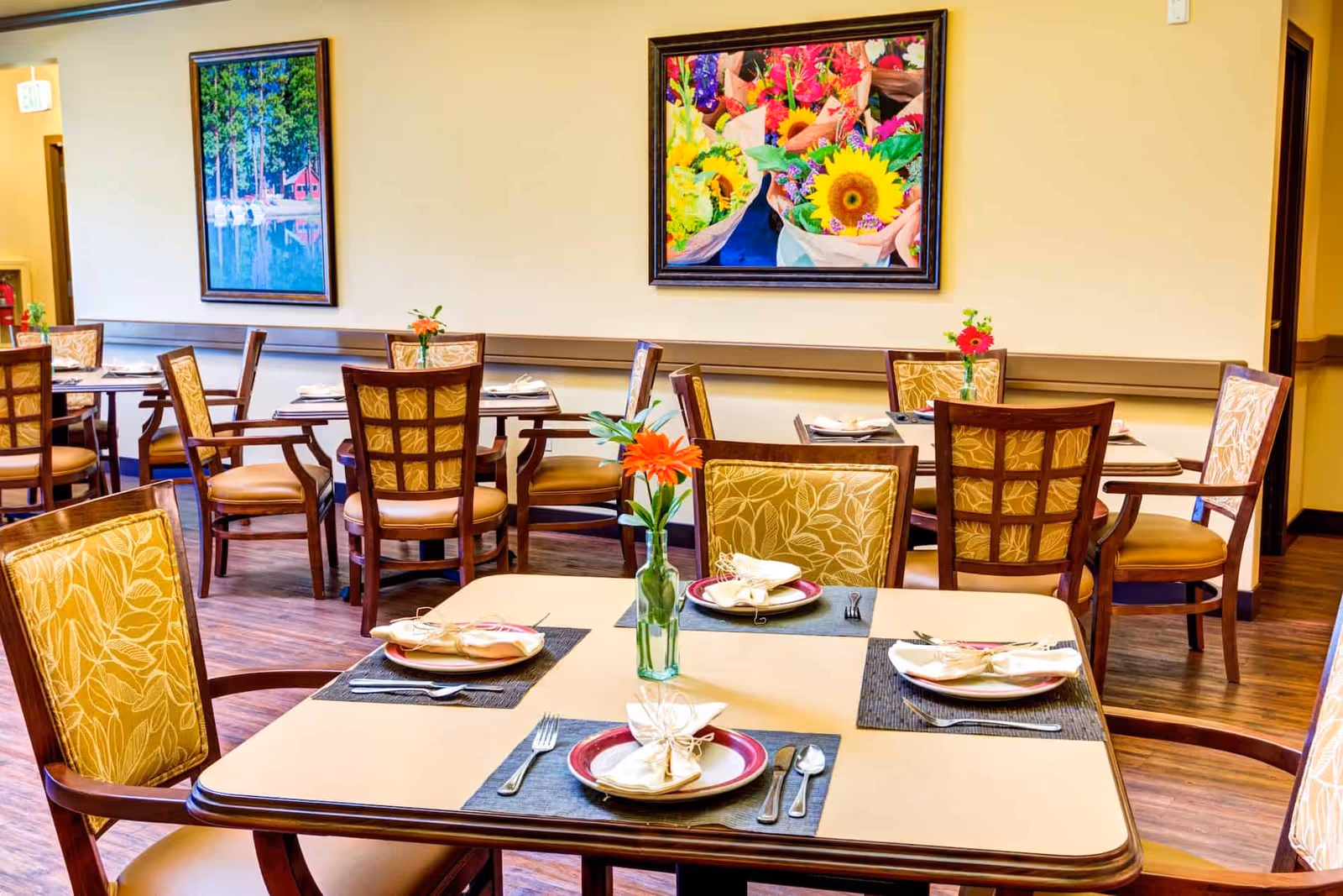 A bright dining room with several tables set for meals, each with plates, napkins, and silverware. The chairs have patterned yellow upholstery. There are colorful flower arrangements in small vases on each table. Two framed paintings hang on the wall, one depicting a lakeside scene and the other a vibrant bouquet of flowers.