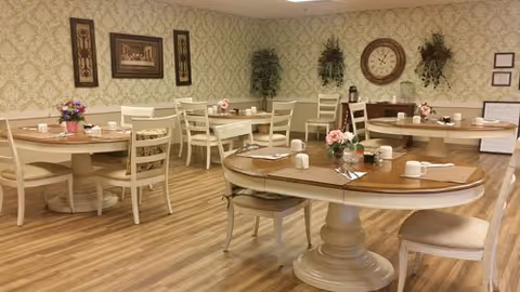 A dining room with several round and rectangular tables set with placemats, cups, and small flower arrangements. The room has light wood flooring, patterned wallpaper, framed artwork, a large wall clock, and decorative plants on the walls.