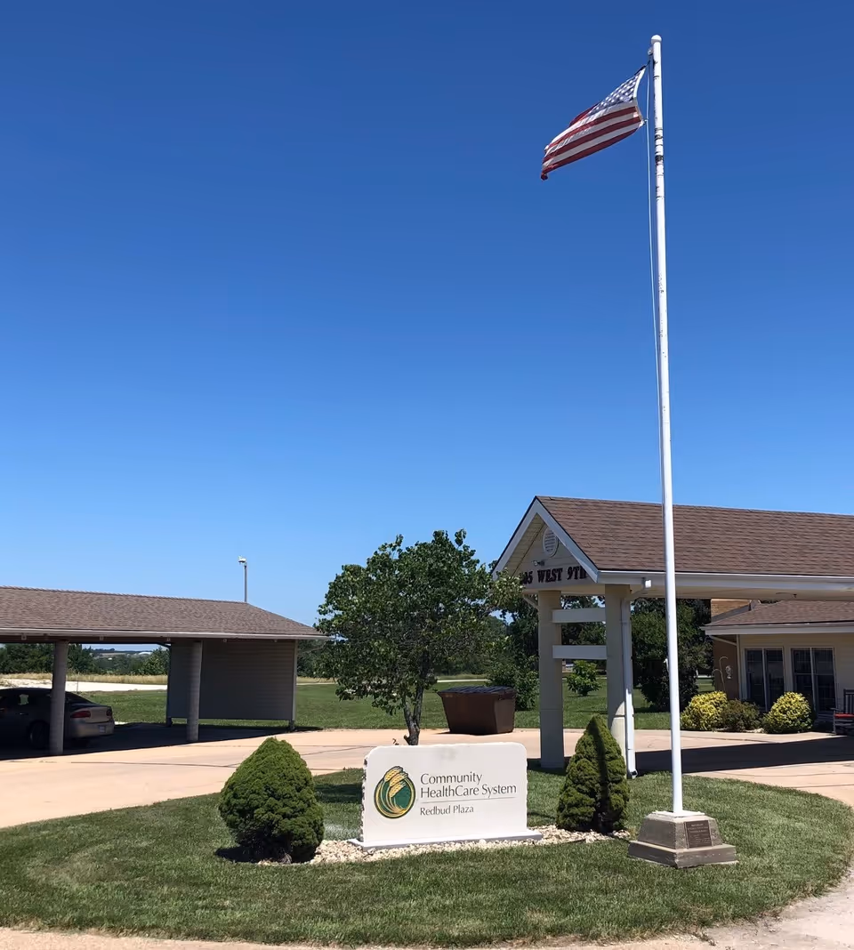 Exterior view of Redbud Plaza Assisted Living facility with a clear blue sky, an American flag on a tall flagpole, a sign reading 'Community HealthCare System Redbud Plaza', a small tree, two small bushes, a covered parking area with a car, and part of the building with windows and shrubs.