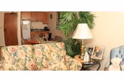 Cozy living area with a floral patterned sofa, a side table holding a lamp and framed photos, a large green plant, and a glimpse into a kitchen area with wooden cabinets and appliances.
