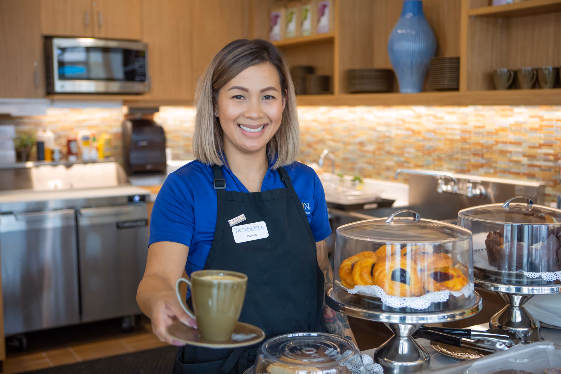 A smiling woman wearing a blue shirt and black apron stands behind a counter in a kitchen area, holding a cup of coffee on a saucer. On the counter in front of her are covered trays with pastries and muffins. The background shows kitchen cabinets, a microwave, and a tiled backsplash.