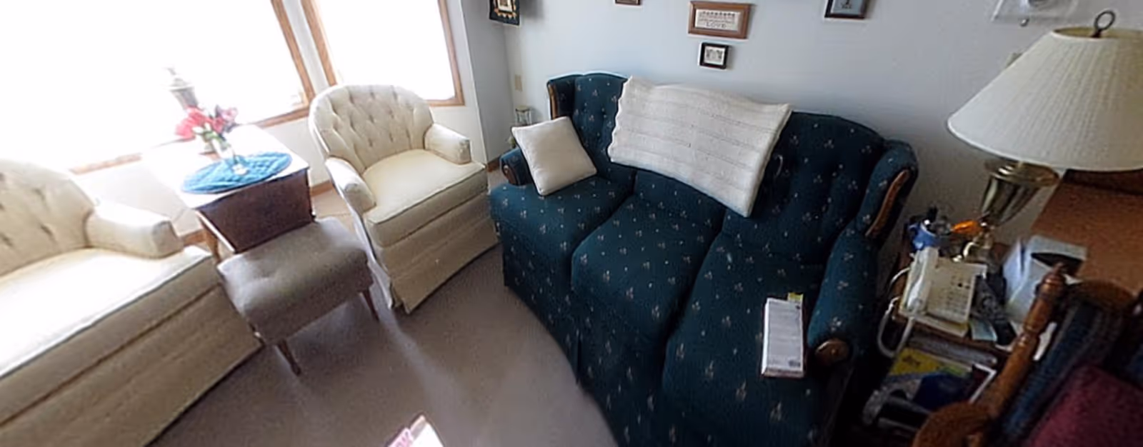 A cozy living room area with a dark green patterned sofa adorned with a white throw blanket and pillow, two cream-colored armchairs, a small ottoman, a wooden side table with a lamp and flowers, and a telephone on a small stand. The room has light-colored walls with framed pictures and a window letting in natural light.