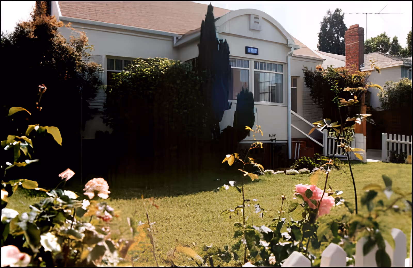 A garden area with green grass and blooming flowers in front of a white house with a small porch and stairs. There are bushes and trees surrounding the house, and a white picket fence is partially visible in the foreground.