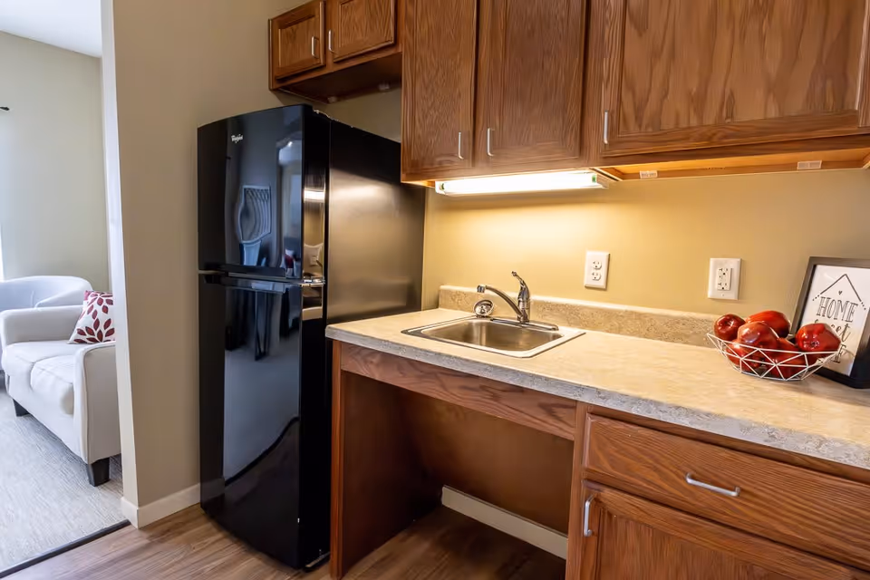 A small kitchen area with wooden cabinets, a black Whirlpool refrigerator, a countertop with a sink, and a bowl of red apples. A framed sign that says 'Home sweet home' is placed on the counter. In the background, there is a white armchair with a red and white patterned pillow.