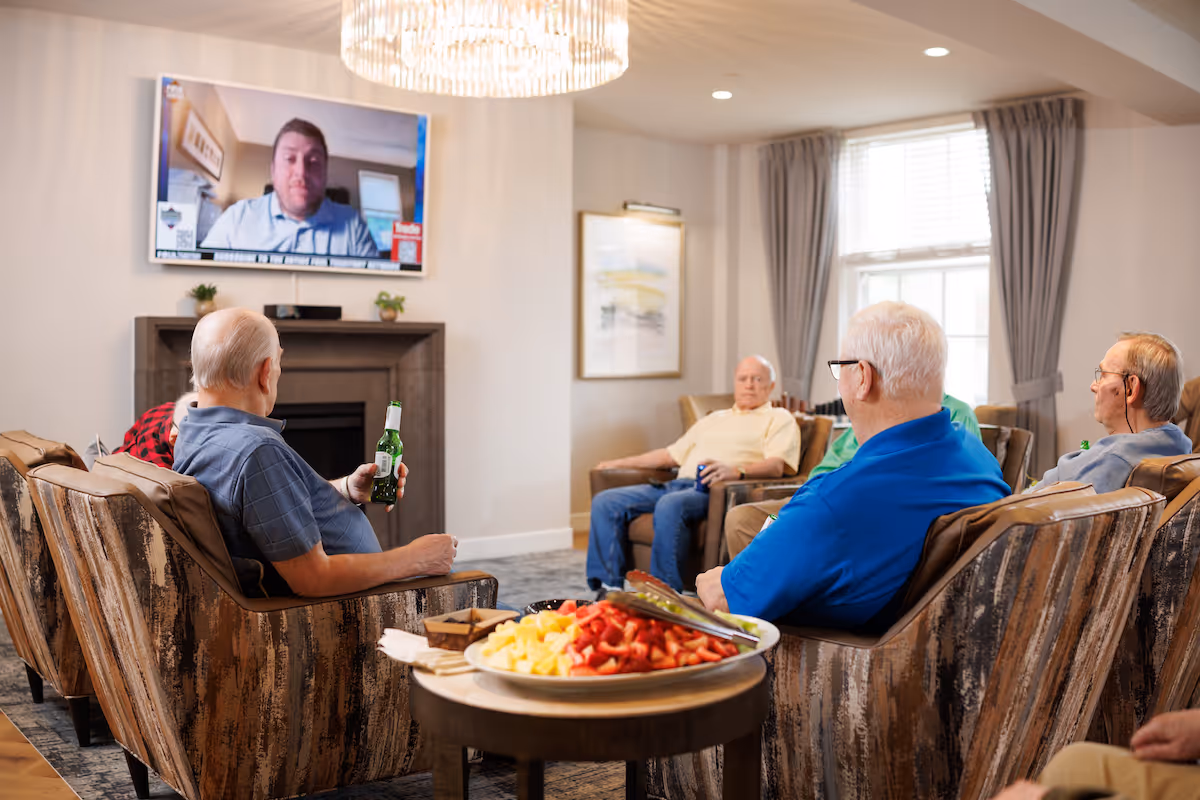Several elderly men seated in a cozy common living room watching a wall-mounted TV while a table with fruit sits in the foreground.