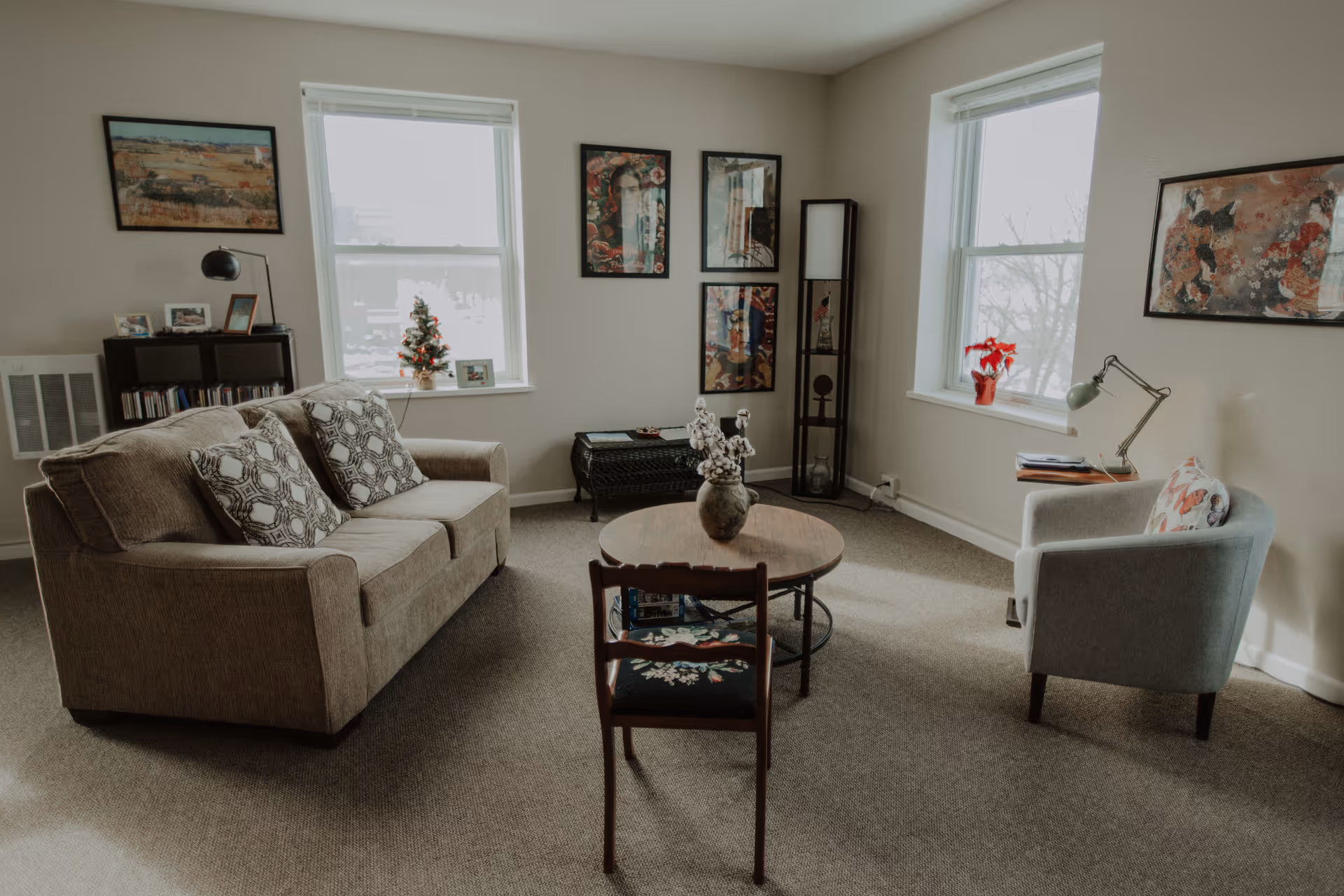 A cozy living room with a beige sofa adorned with patterned pillows, a wooden chair with floral upholstery, and a light gray armchair with a butterfly pillow. The room has two windows with white blinds, framed artwork on the walls, a small decorated Christmas tree on the windowsill, a red poinsettia plant on another windowsill, a round wooden coffee table with a vase of cotton stems, and a black shelving unit with photos and books.