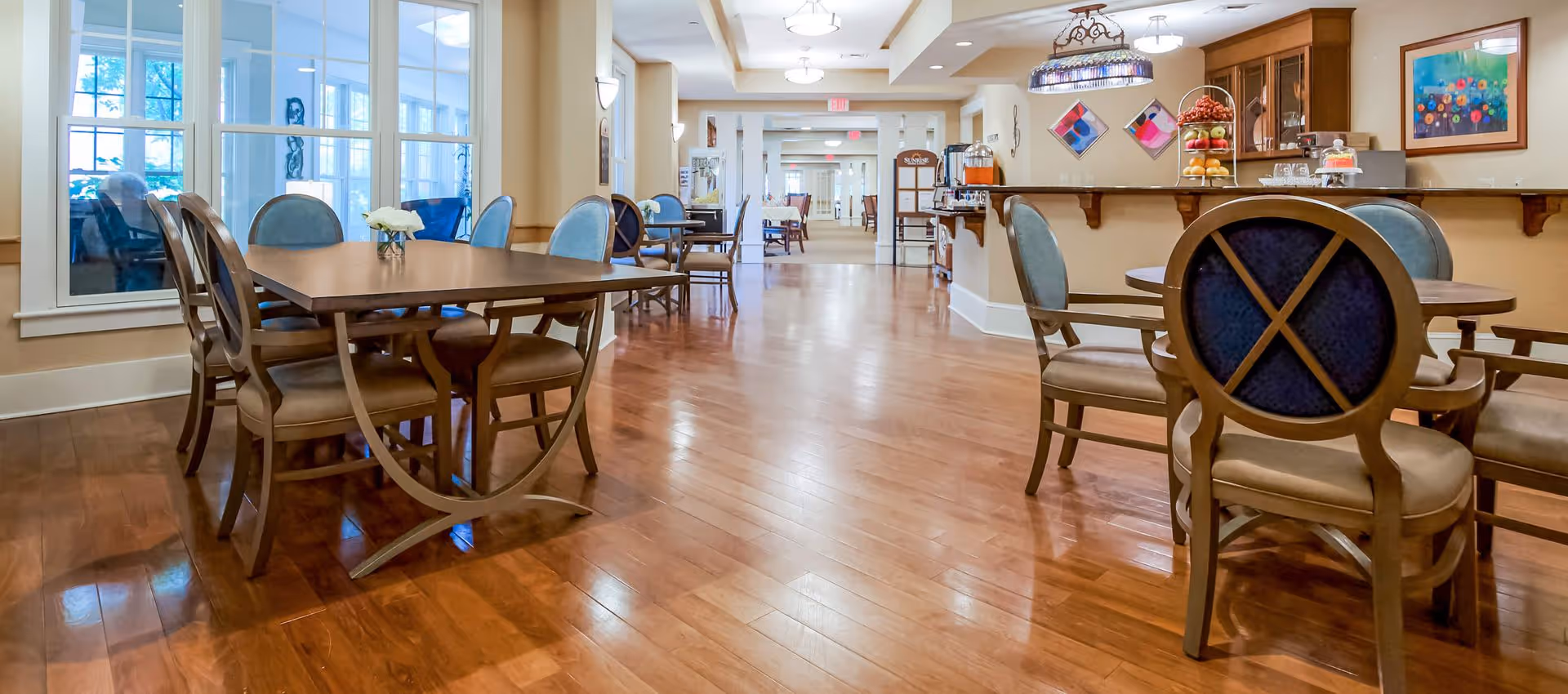 A spacious dining area with wooden floors, multiple wooden tables and chairs with blue cushioned seats, a counter with fruit and a cake on display, and colorful artwork on the walls. Large windows on the left side let in natural light.