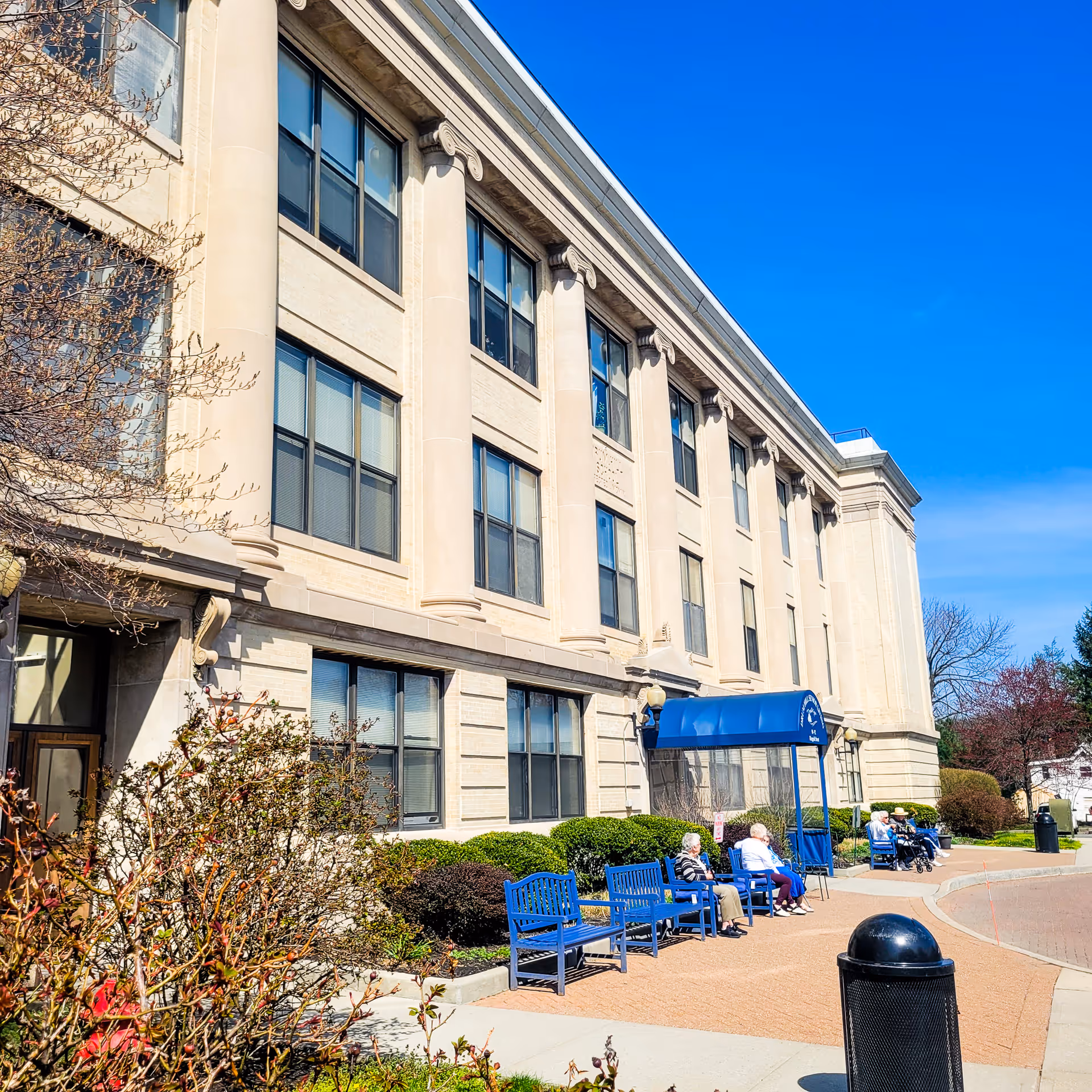 Front exterior of a multi-story senior living building with a blue entrance awning and benches where residents sit.